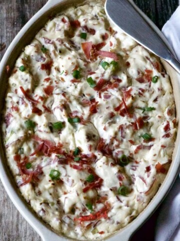 A baked dish with a creamy, white topping, garnished with chopped green onions and red bits of meat, placed on a wooden surface with a silver spoon on the side.