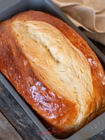A loaf of freshly baked paska bread with a golden crust sits in a metal baking pan on a wooden surface, next to a beige cloth.