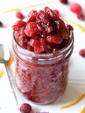 A glass jar filled with cranberry sauce sits on a white surface, surrounded by loose cranberries and orange zest, with a spoon beside it-an inviting scene reminiscent of homemade Cranberry Apple Chutney.
